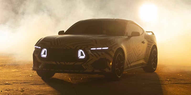 Three-quarter front view of a camouflaged sports coupe parked on a dusty surface at sunset