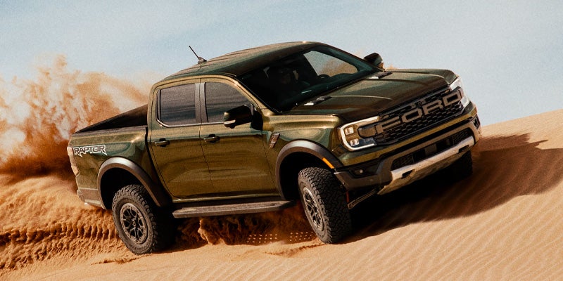 Dark green off-road pickup truck driving across sand dunes, kicking up sand while descending a steep slope