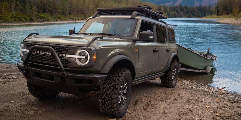 Gray off-road SUV parked beside a river with a small boat attached, surrounded by forested mountains and rocky shoreline
