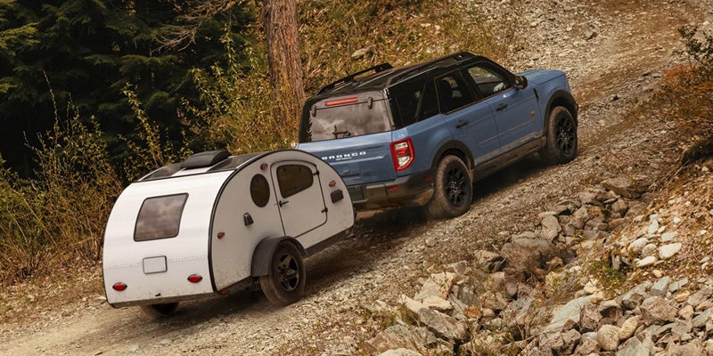 Blue SUV towing a compact white travel trailer up a steep, rocky dirt road through a forested mountain landscape.