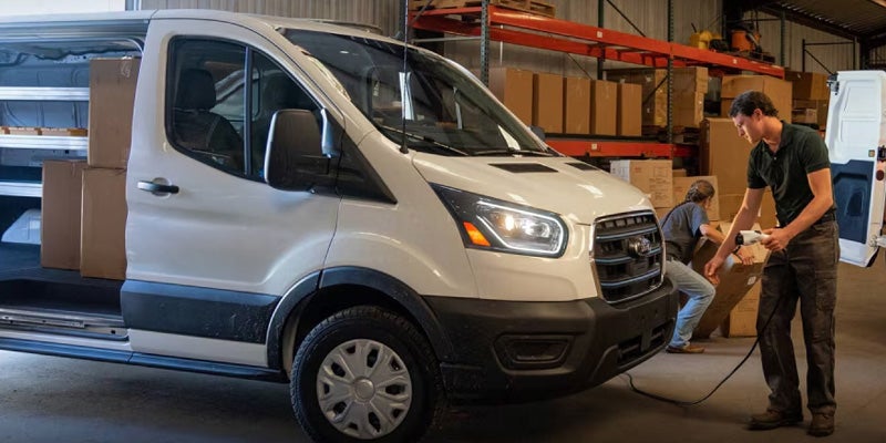 White electric van being charged while workers load boxes in a warehouse.