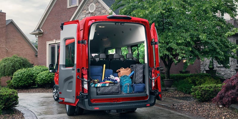 Red van with rear doors open, cargo area packed with luggage, parked in a driveway in front of a house.