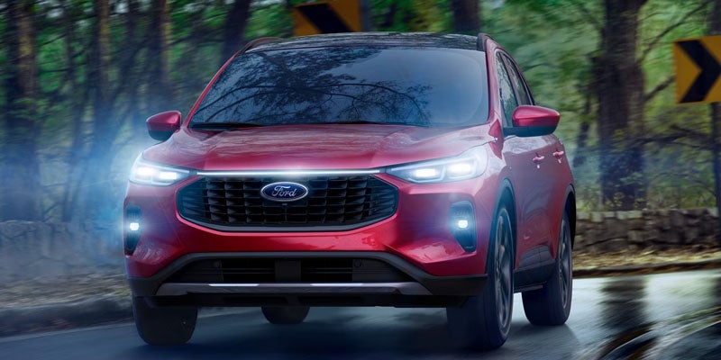 A red 2024 Ford Escape driving on a wet, tree-lined road with its headlights and fog lights illuminated.