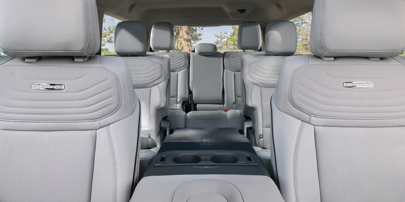 Light gray car interior with two rows of quilted leather seats, center console with cupholders, and a view of trees outside.