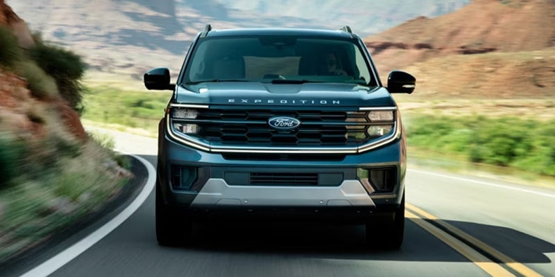 Front view of a dark blue Ford Expedition SUV driving on a winding road with rocky hills in the background.