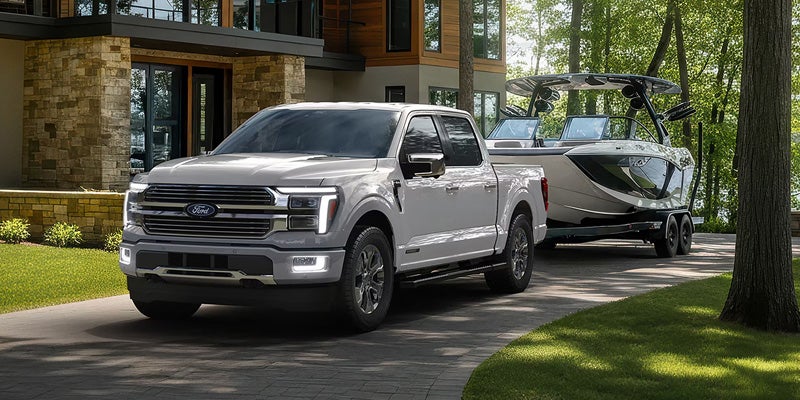 A light gray Ford F-150 truck with LED headlights on, towing a silver and white boat on a trailer, parked on a paved driveway in front of a modern house.