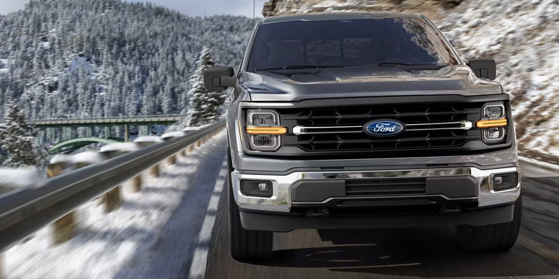 A gray Ford F-150 pickup truck drives on a snowy road with pine trees and a bridge in the background.