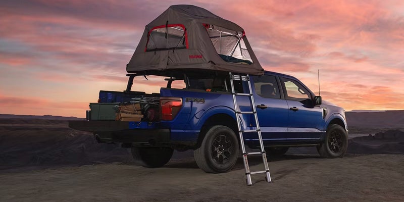 Blue 2026 Ford F-150 with rooftop tent and camping gear at sunset.