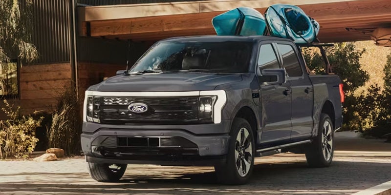 Dark grey Ford F-150 Lightning pickup truck with two blue kayaks on the roof rack, parked in front of a modern home.
